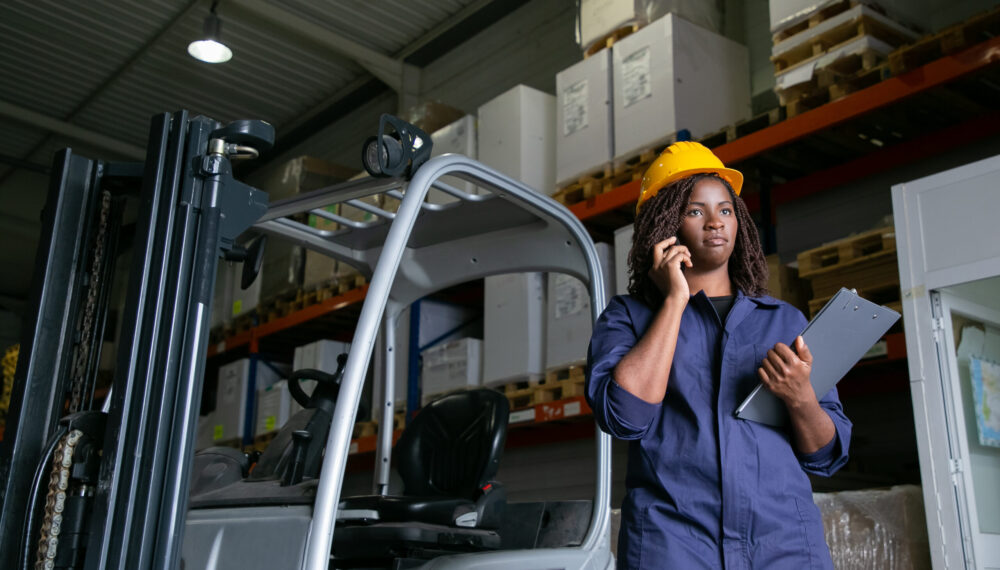 Serious female warehouse employee in hardhat standing near forklift and talking on mobile phone. Shelves with goods in background. Copy space. Labor or communication concept