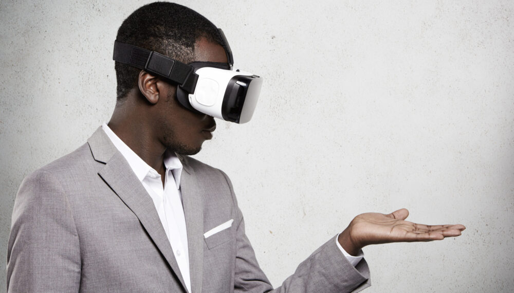 Technology, entertainment, gaming and cyberspace. African office worker in formal suit experiencing virtual reality using 3d headset for mobile phone, looking at his open palm as if holding something