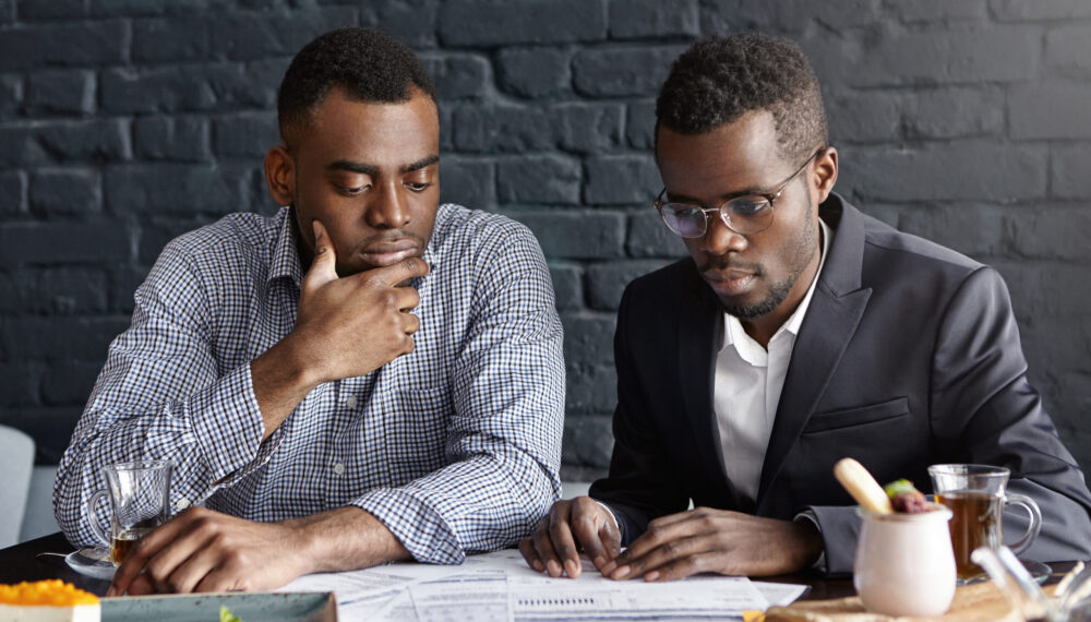 Two handsome dark-skinned executives having thoughtful and serious facial expression while reviewing finances and doing paperwork together, sitting at desk with documents in modern office interior