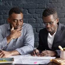 Two handsome dark-skinned executives having thoughtful and serious facial expression while reviewing finances and doing paperwork together, sitting at desk with documents in modern office interior